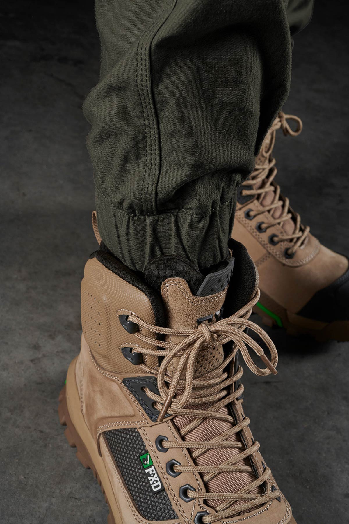A person wears FXD WP-4 Stretch Cuffed Work Pants tucked into tan hiking boots with thick soles and detailed stitching, standing on a dark textured floor.