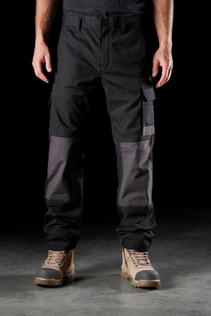 A person stands facing forward against a black background, wearing FXD WP-1 black cargo pants with Duratech reinforced pockets and 360-degree stretch for maximum flexibility, black clothing on top, and tan work boots with dark laces and green soles. The image is cropped from the shoulders down.