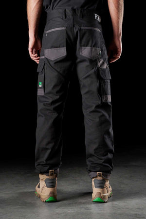 A person wearing FXD WP-1 black, multi-pocket work pants stands on a gray surface, facing away from the camera. They are also wearing beige work boots with green soles. The comfortable FXD WP-1 pants feature gray patch pockets and Duratech reinforced pockets, with the "FXD" logo visible on the waistband.