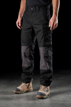 A person stands on a concrete surface wearing FXD WP-1 work pants with Duratech reinforced pockets and tan work boots. The background is dark, highlighting the pants and boots. Only the lower half of the person is visible.