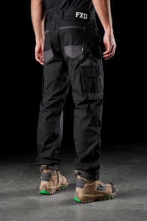 A person stands with their back facing the camera, wearing FXD WP-1 black comfortable work pants featuring Duratech reinforced pockets and "FXD" written on the back. They are also wearing tan and black work boots with green accents.