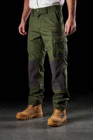 A person wearing FXD WP-1 green cargo work pants with black knee patches, a white shirt, and tan work boots. The FXD WP-1 pants offer 360-degree stretch and feature multiple pockets, including a visible logo on the right thigh and Duratech reinforced pockets. The plain dark background emphasizes the clothing and footwear.