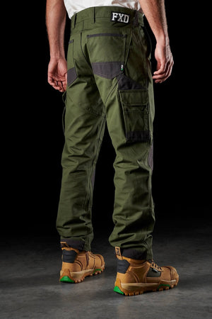 An individual clad in FXD WP-1 green cargo work pants, featuring Duratech reinforced pockets and black patch details on the knees, stands with their back to the camera. They are also sporting brown work boots accented with orange and green soles. The backdrop is black.