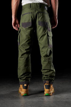 A person stands with their back to the camera, showcasing FXD WP-1 dark green utility work pants with Duratech reinforced pockets and brown work boots. The background is black, emphasizing the durable design of both the pants and boots.