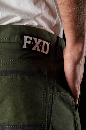 Close-up of a person wearing FXD WP-1 green work pants and a white shirt. The focus is on the back pocket area with a black and white "FXD" label on the waistband. A portion of the person's hand is visible, resting casually on the side, highlighting the Duratech reinforced pockets for enhanced durability.