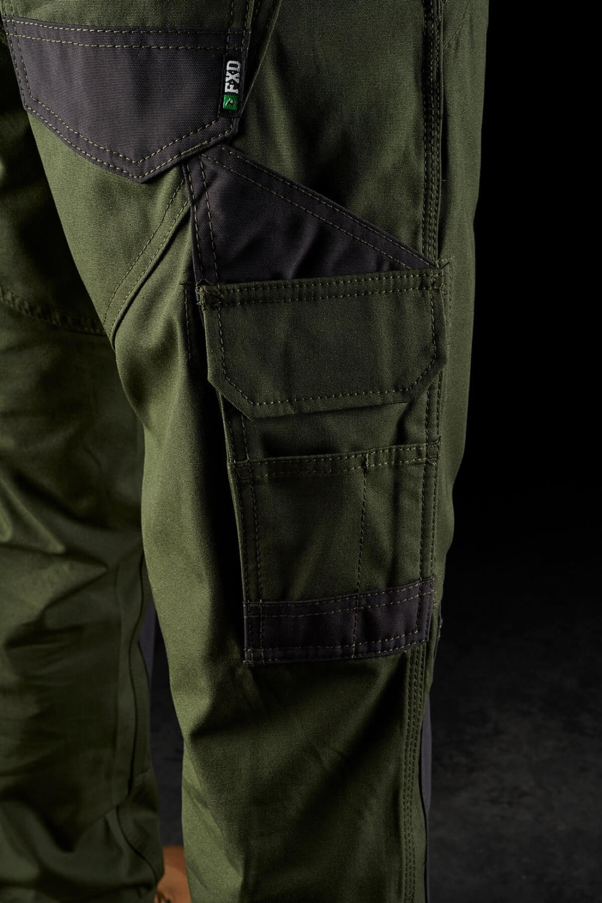Close-up of a person wearing FXD WP-1 green work pants with dark patches and multiple Duratech reinforced pockets. The FXD WP-1 pants, featuring 360-degree stretch, have a rugged and tactical look suitable for outdoor or heavy-duty activities. The background is dark, highlighting the pants' details and texture.