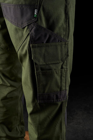 Close-up of a person wearing FXD WP-1 green work pants with dark patches and multiple Duratech reinforced pockets. The FXD WP-1 pants, featuring 360-degree stretch, have a rugged and tactical look suitable for outdoor or heavy-duty activities. The background is dark, highlighting the pants' details and texture.