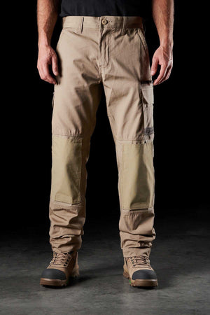 A person wearing the FXD WP-1 beige cargo pants with Duratech reinforced pockets and a black shirt stands against a black background. They are also donning brown work boots. Designed as comfortable work pants, the FXD WP-1 features multiple pockets and offers 360-degree stretch for durability.