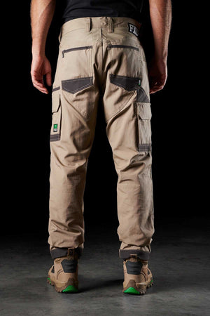 A person stands facing away, wearing the FXD WP-1 beige utility trousers with multiple pockets and black accents. Featuring Duratech reinforced pockets and 360-degree stretch for maximum flexibility, these comfortable work pants have a brand logo on the waistband and side pocket. The person is also wearing brown and green work boots against a dark background.