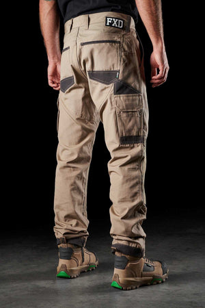 A person is standing with their back to the camera, wearing FXD WP-1 light brown comfortable work pants featuring multiple Duratech reinforced pockets and black patches. The person also has a black shirt and brown work boots with green soles. The setting is a dark background with a concrete floor.