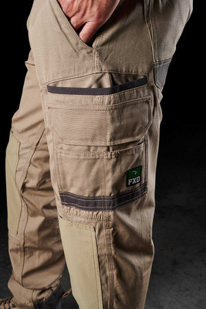 Close-up image of a person wearing FXD WP-1 beige work pants featuring multiple utility pockets, including one with a small black and green FXD logo patch. The Duratech reinforced pockets ensure durability, while the 360-degree stretch fabric offers ultimate comfort. The person's hand is partially visible resting on one of the pockets. The background is dark.