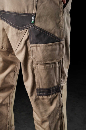 Close-up of the lower half of a person wearing FXD WP-1 tan work pants, featuring multiple pockets with black reinforced stitching and showcasing the Duratech reinforced pockets. A small green and white label is attached near one of the pockets, highlighting the comfortable design. The background is black.