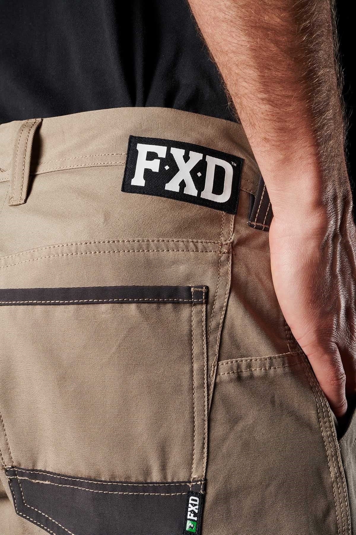 Close-up of a person wearing FXD WP-1 work pants in beige, showcasing visible stitching and Duratech reinforced pockets. The person is standing with their right arm resting by their side. The FXD logo is prominently displayed on the waistband and pocket label.
