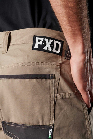 Close-up of a person wearing FXD WP-1 work pants in beige, showcasing visible stitching and Duratech reinforced pockets. The person is standing with their right arm resting by their side. The FXD logo is prominently displayed on the waistband and pocket label.