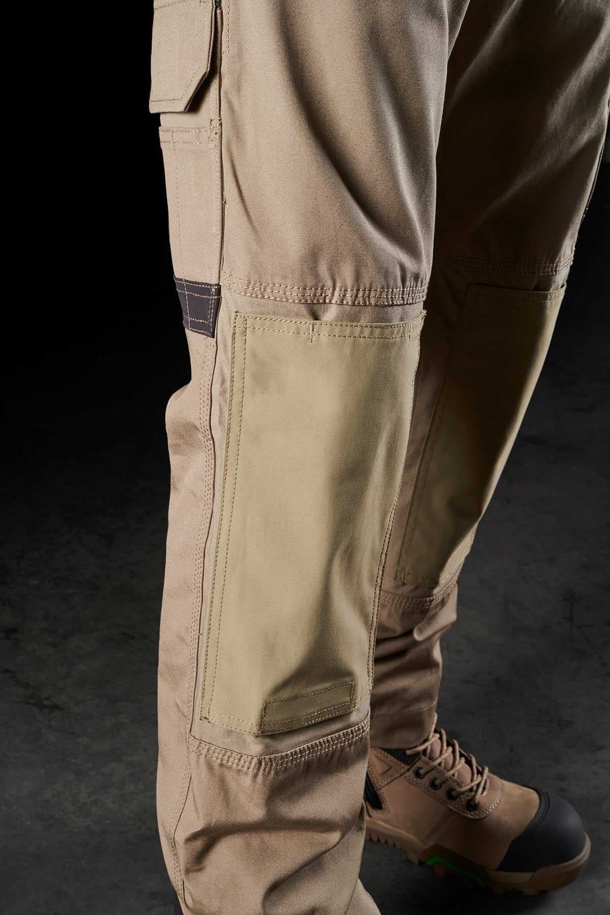 Close-up of a person wearing FXD WP-1 tan cargo pants with reinforced knee patches and brown work boots. The comfortable FXD WP-1 features Duratech reinforced pockets and a side pocket with a flap. The boots, adorned with green accents, complement the rugged attire set against a dark background.