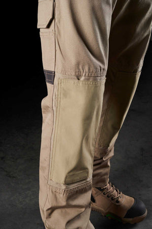 Close-up of a person wearing FXD WP-1 tan cargo pants with reinforced knee patches and brown work boots. The comfortable FXD WP-1 features Duratech reinforced pockets and a side pocket with a flap. The boots, adorned with green accents, complement the rugged attire set against a dark background.