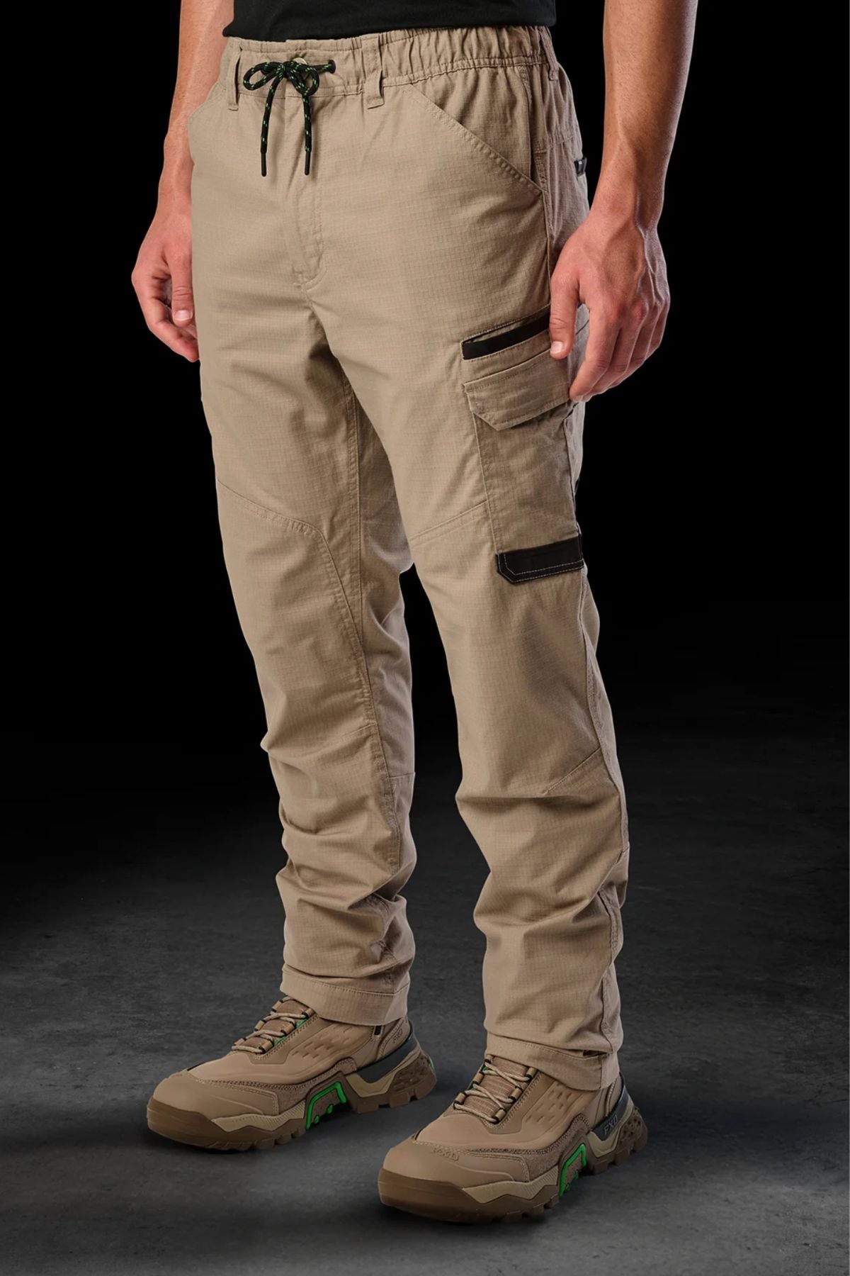A person in FXD WP-12 tan 2-way stretch ripstop pants with black zippers, utility pockets, and a drawstring waist, paired with beige hiking shoes, stands against a dark background. Only the lower half is visible.