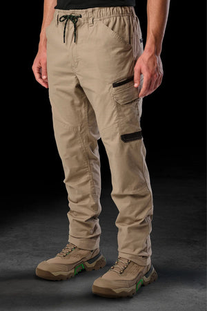A person in FXD WP-12 tan 2-way stretch ripstop pants with black zippers, utility pockets, and a drawstring waist, paired with beige hiking shoes, stands against a dark background. Only the lower half is visible.