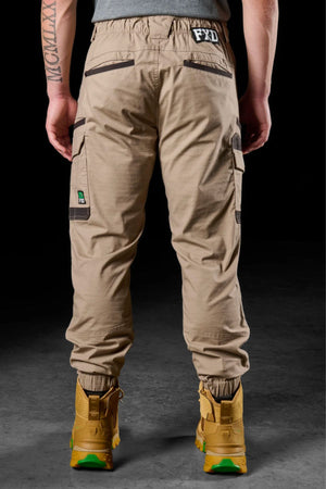 A person, seen from behind on a dark surface, is wearing FXD WP-12C stretch ripstop work pants with multiple pockets and yellow work boots, showing a tattoo on their left forearm.