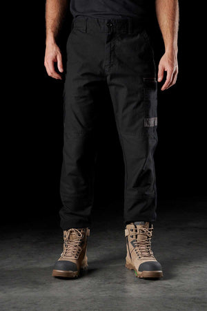 A person wearing FXD WP-3 black stretch cotton cargo pants with reinforced pockets and tan lace-up combat boots stands against a dark background. The FXD WP-3 trousers have side pockets, and the boots feature robust soles and detailing. The person's upper body is not visible.