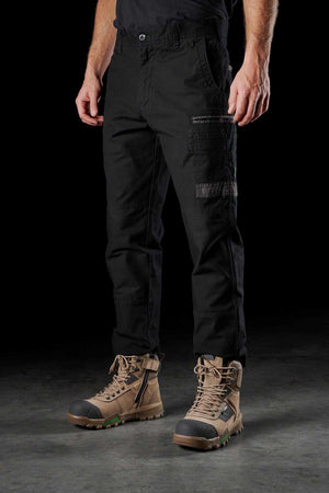 A person wearing FXD WP-3 black stretch cotton cargo pants with reinforced pockets and brown lace-up work boots stands against a dark background. The image focuses on the lower half of their body, showcasing the detailed design of the FXD WP-3 work pant and the rugged, sturdy boots.