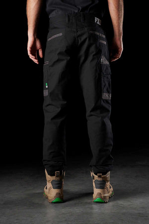 A person stands facing away, sporting the FXD WP-3 black utility work pants with reinforced pockets and brown hiking boots featuring green-accented soles. The dark background makes the outfit stand out clearly.