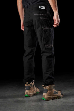 A person wearing FXD WP-3 black cargo pants with reinforced pockets, a black shirt, and tan hiking boots with green accents stands on a dark surface. The image is taken from behind, highlighting the back view of the work pants and boots.