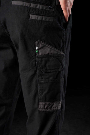 Close-up of the side of a person wearing FXD WP-3 black stretch cotton work pants featuring multiple reinforced pockets. The person has their hand in one of the pockets, and the pants have labels with green and white lettering. The background is dark.