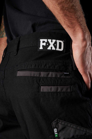 Close-up of a person wearing FXD WP-3 black work pants. The stretch cotton fabric provides both comfort and flexibility, while the reinforced pockets with zippers and intricate stitching details guarantee durability. The model's right hand rests on their hip, highlighting the back pocket area and prominent FXD logo against a dark background.