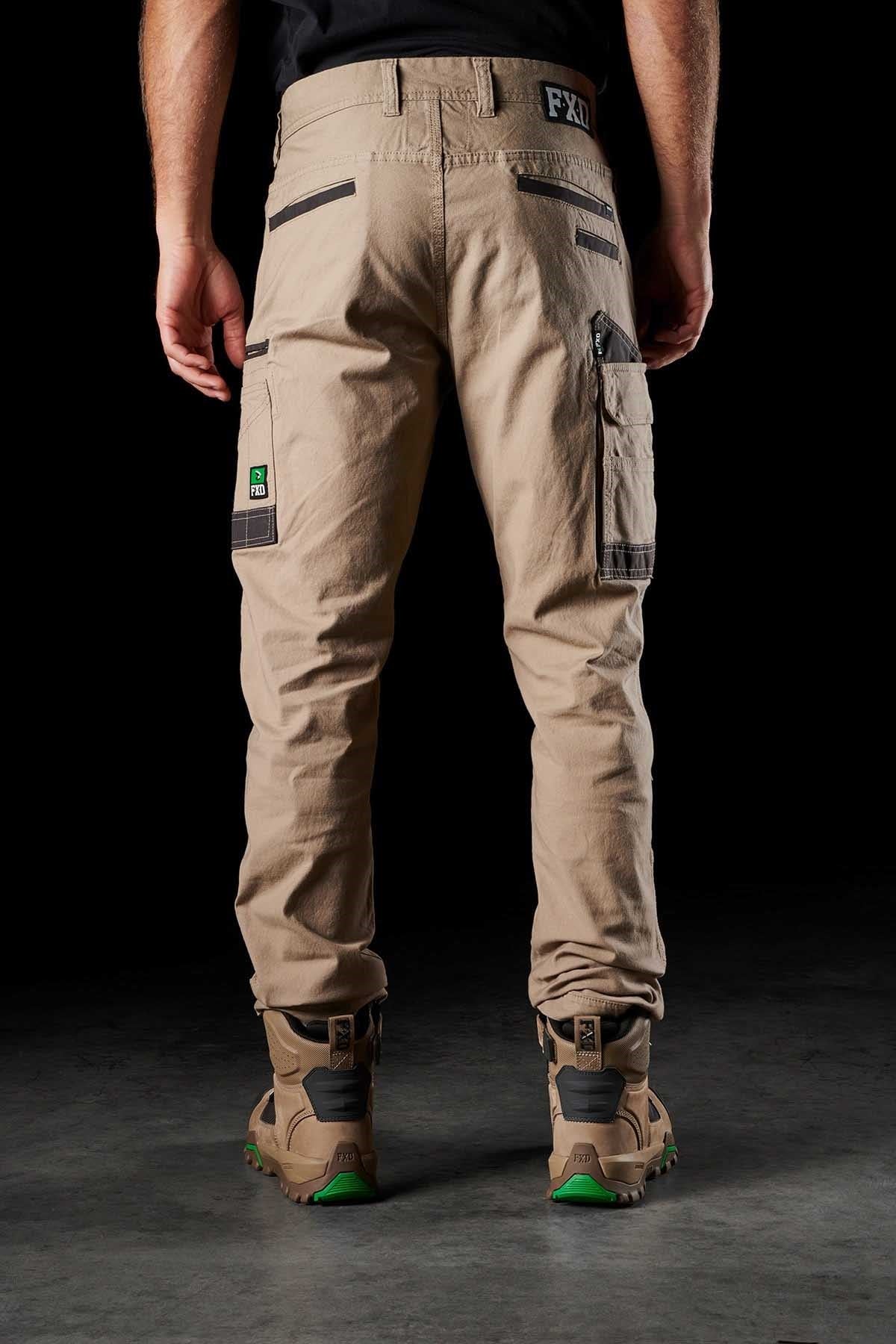 A person stands facing away, wearing the FXD WP-3 tan utility pants with reinforced pockets and black zippers, paired with work boots featuring green accents. The background is completely black, highlighting the details of the outfit.