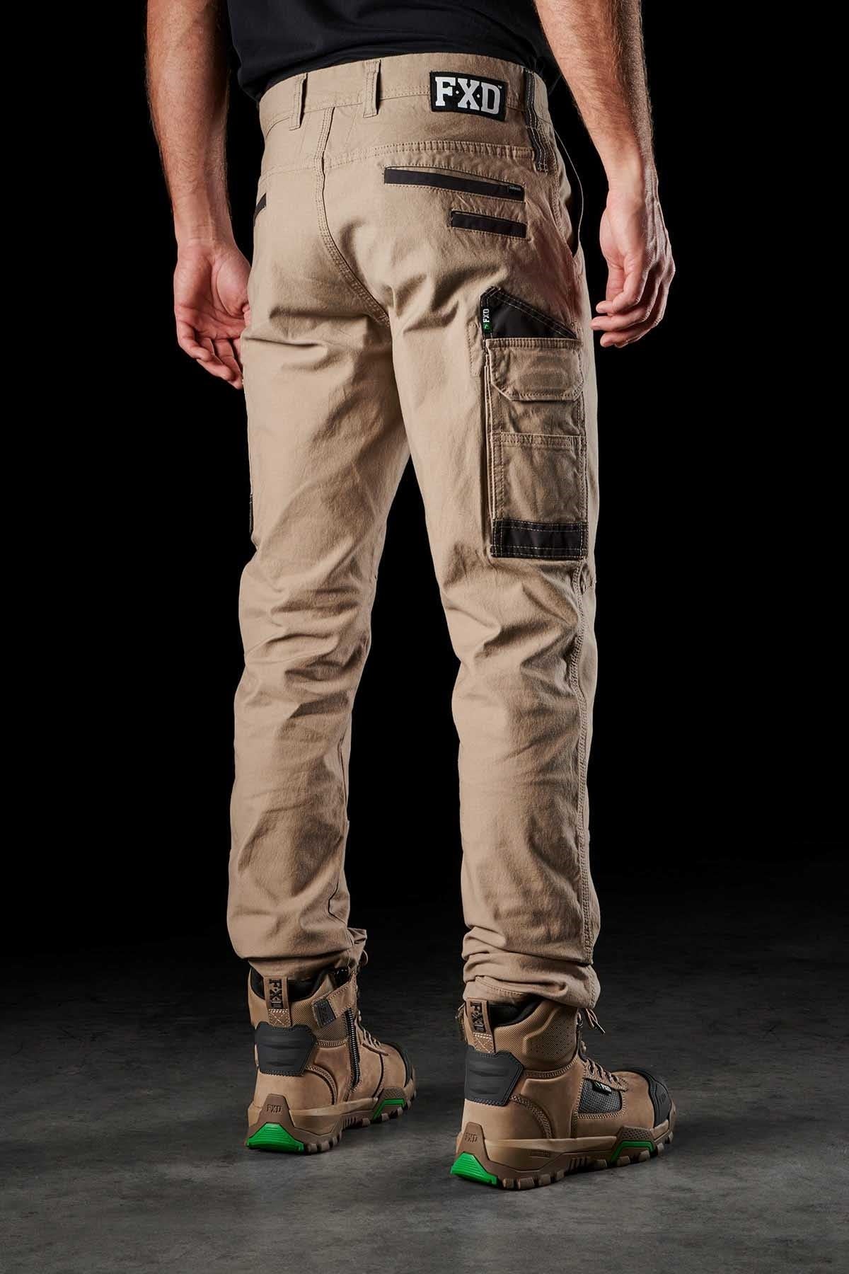 A person stands facing away, wearing FXD WP-3 beige cargo pants with black detailing, reinforced pockets, and "FXD" printed on the waistband. They are also wearing brown and green work boots. The solid black background highlights the outfit.
