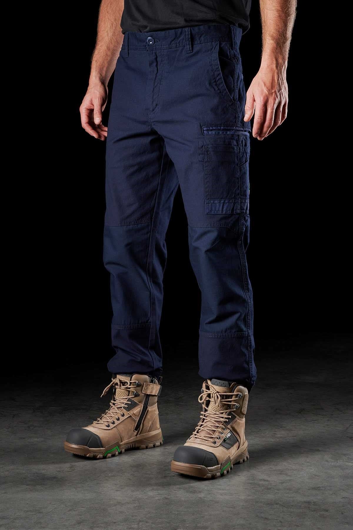 Against a dark background, a person stands wearing FXD WP-3 navy blue cotton cargo pants with multiple pockets and rugged, tan lace-up work boots. The individual is also sporting a black shirt, though only the lower torso and legs are visible in the image.