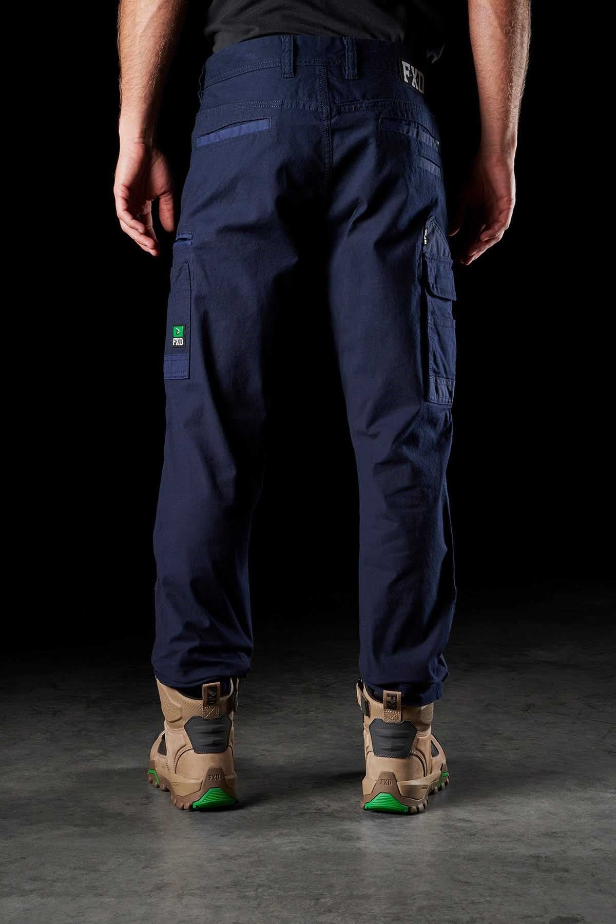 A person is standing in a dim environment, dressed in navy blue cargo pants and tan work boots. The focus is from the waist down, emphasizing the FXD WP-3 Cotton work pant that provides 360-degree stretch. Their hands are resting on the sides of their legs.