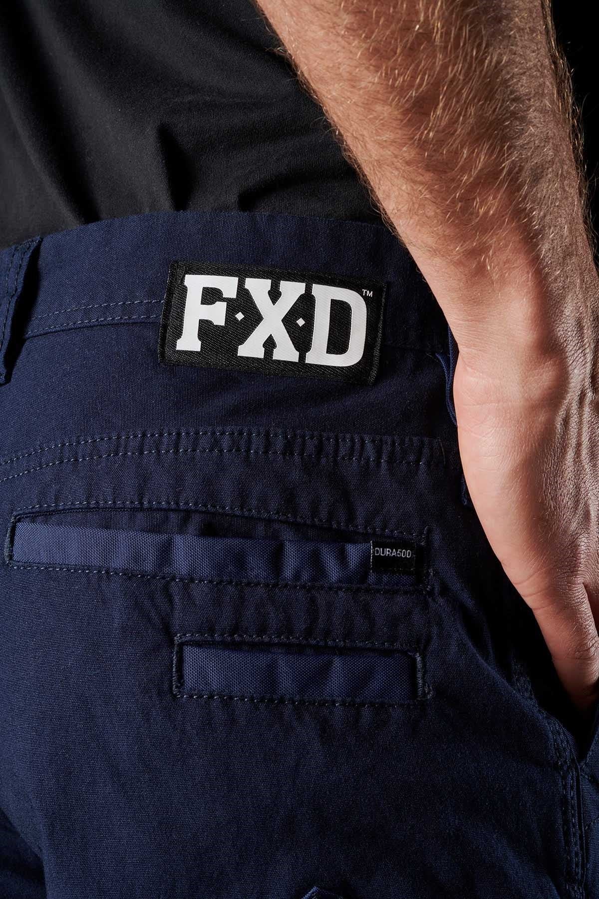 Close-up view of a person wearing FXD WP-3 work pants in dark blue, showcasing an FXD logo patch on the back waistband and a DURASCO label near the rear pocket. Made from Formatik Cotton, these work pants provide 360-degree stretch for optimal comfort and flexibility. The person's arm rests against their hip in the darkened background.