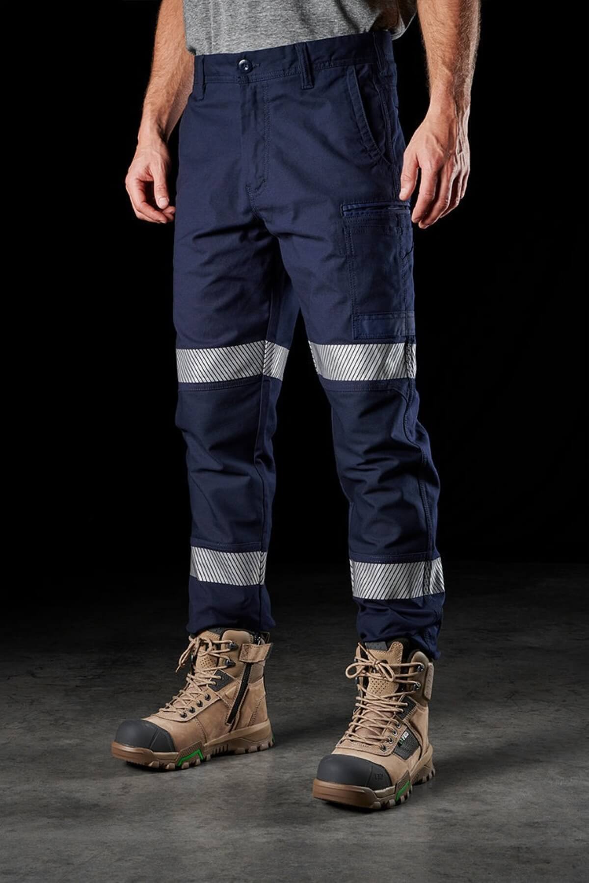 Person wearing FXD WP-3T blue cargo work pants with reflective tape above the knees and sturdy tan boots. The background is dark, highlighting the pants and boots. The person's upper body is mostly out of the frame, with only a portion of a gray shirt visible.
