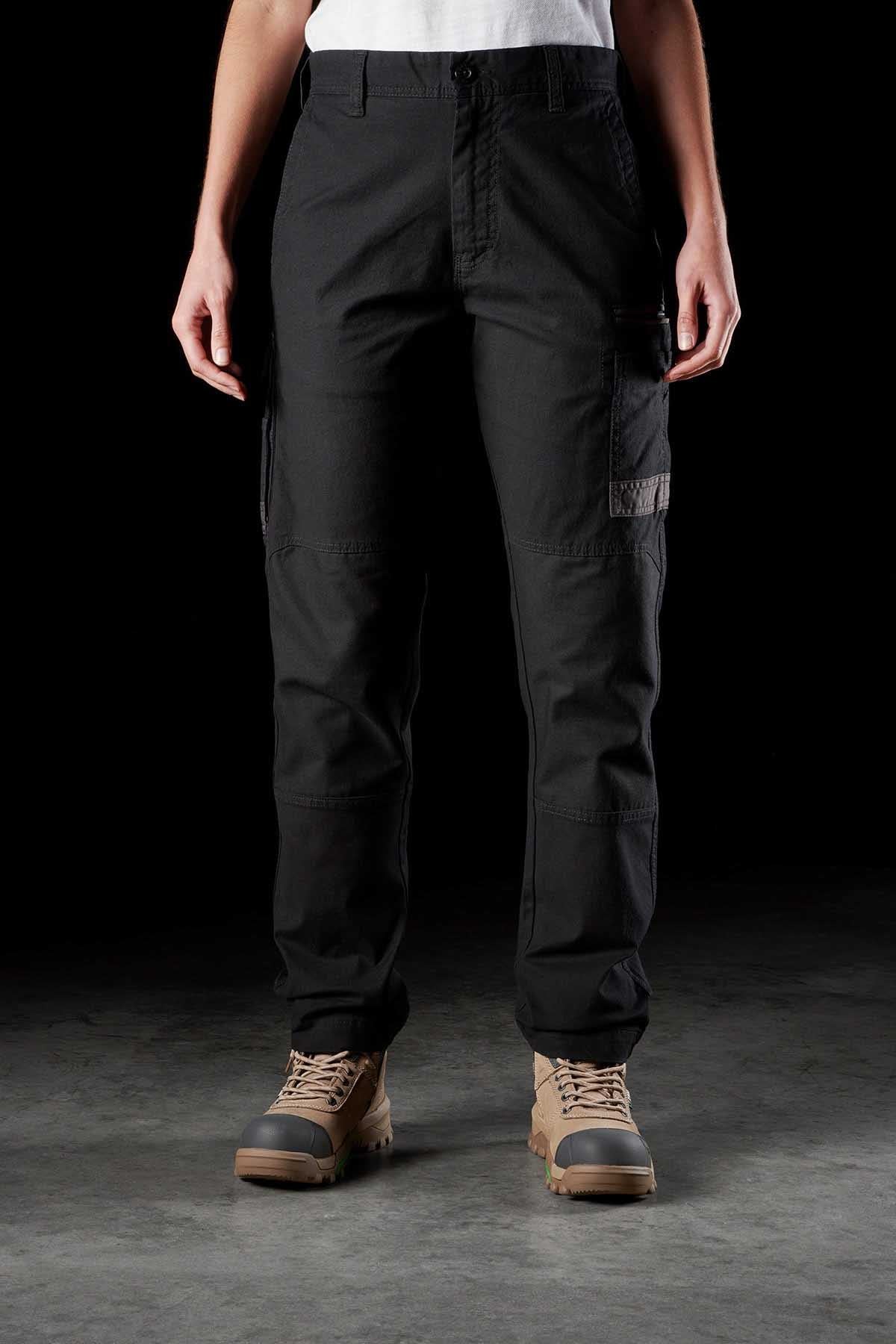 A person stands against a dark background, wearing a white t-shirt, FXD Womens WP-3W black cargo pants with multiple reinforced pockets, and tan lace-up work boots. The photo is focused on their lower half, from the waist to the feet.