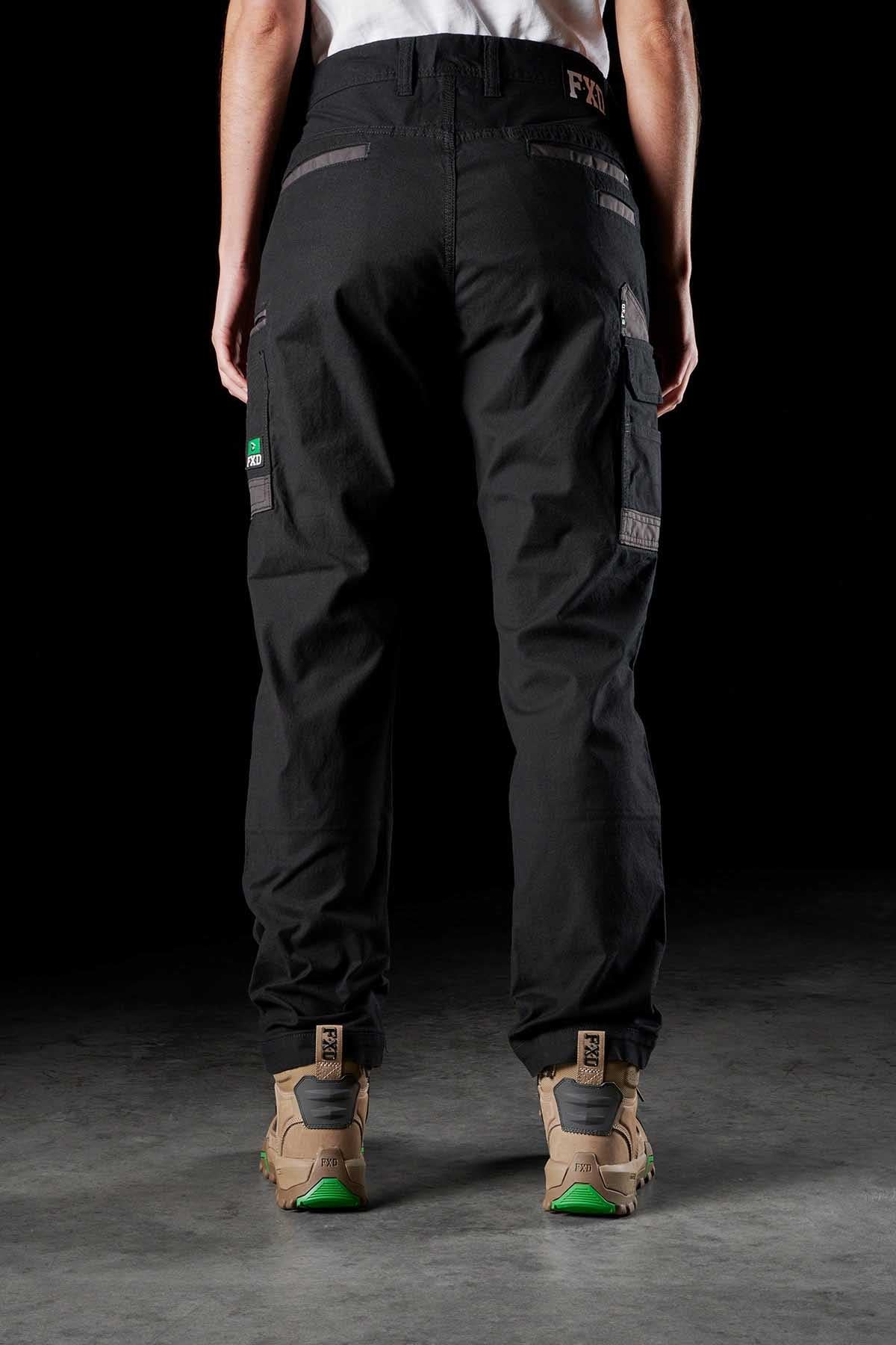An individual is depicted from the back, donning dark FXD Womens WP-3W work pants with reinforced pockets and patches, along with tan hiking boots featuring green soles. The brand logos and details of the women’s work pants and boots are visible. The setting is a dimly lit area with a concrete floor.