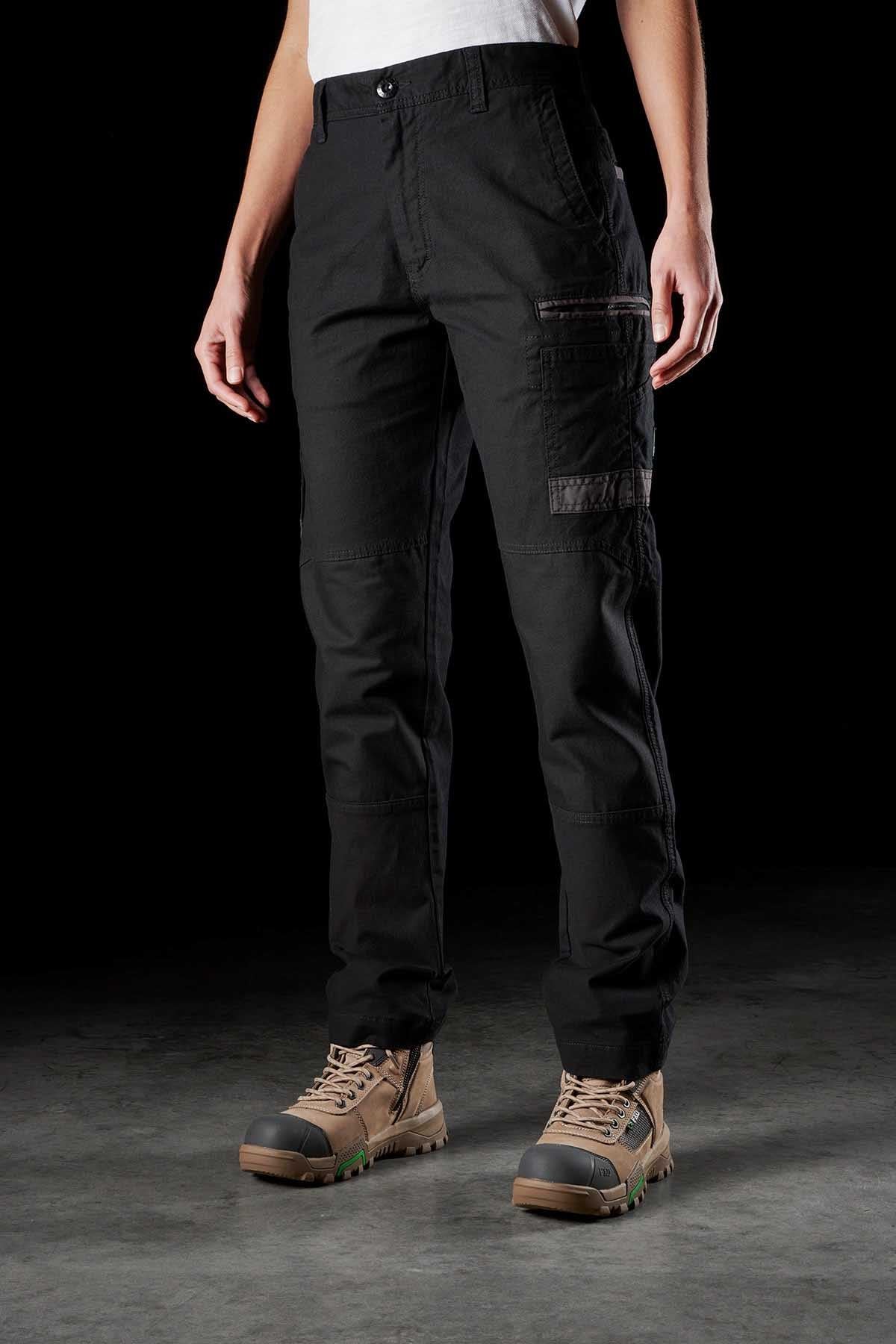 A person wearing FXD Women's WP-3W black cargo pants with multiple reinforced pockets and tan hiking boots stands against a dark background. The work pant, paired with a tucked-in white shirt, offers 360-degree stretch for ultimate comfort. Only the legs and torso are visible, not the head.