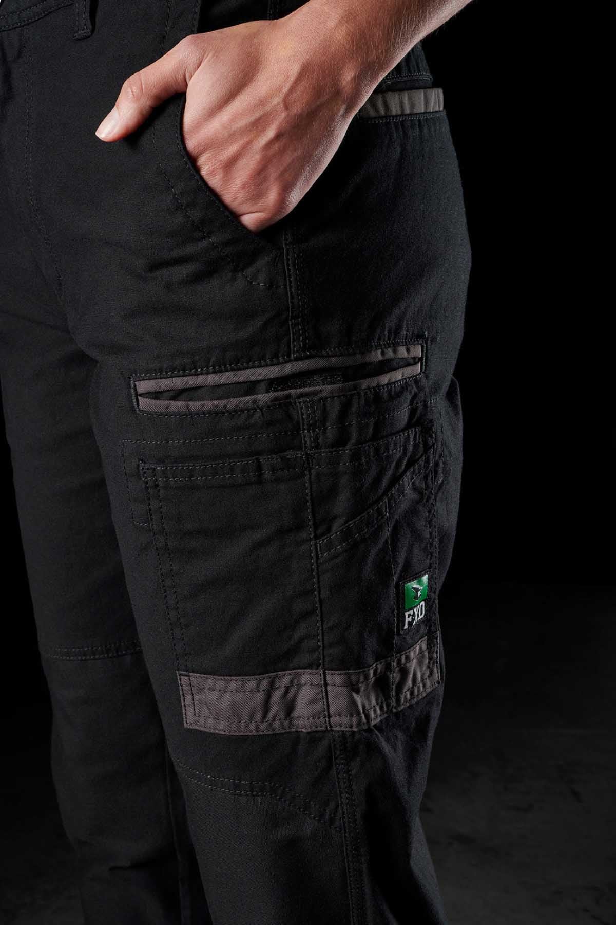 Close-up of a person wearing FXD Womens WP-3W black cargo pants with hands in pockets. The women's work pant features multiple reinforced pockets, a gray reflective stripe, and a small embroidered green and white patch with "FDI" on it. The background is black.