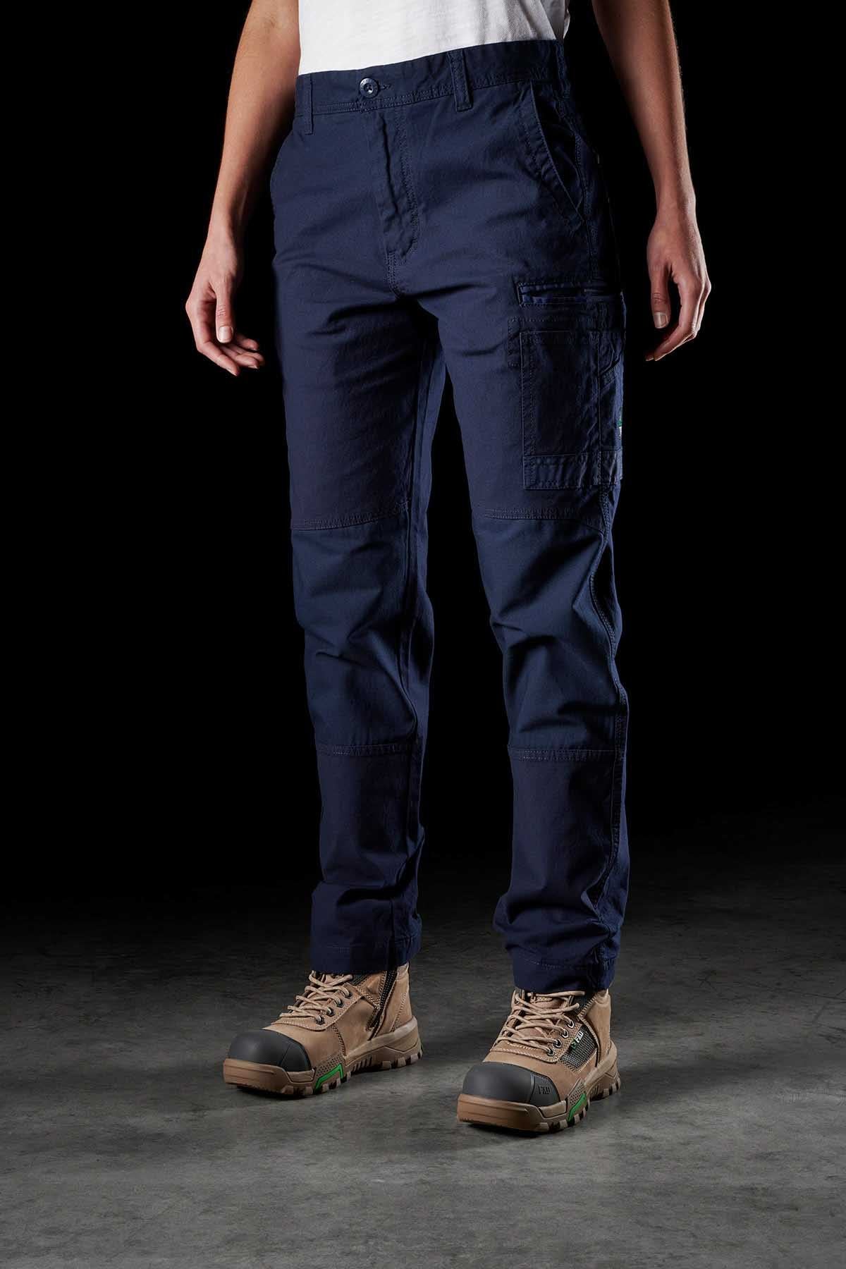 A person stands against a dark background, wearing a white shirt, FXD Womens WP-3W navy blue cargo pants with reinforced pockets, and tan work boots. The cargo pants have multiple pockets visible on the front. Only the person's torso and legs are visible in the image.