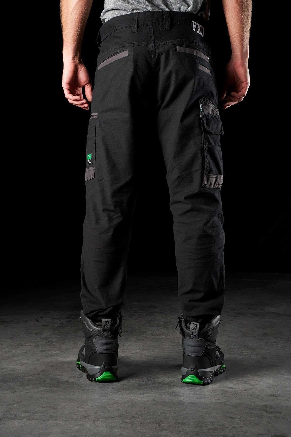 With their back facing the camera, a person is wearing black FXD WP-4 Stretch Cuffed Work Pants featuring pockets, paired with gray and green sneakers. The dark background accentuates the lower half of their body, highlighting the durability of DURA500 Advanced Nylon and stretch cotton canvas material.