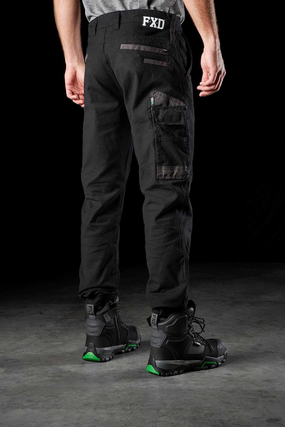 A person is standing, facing away from the camera, wearing black FXD WP-4 Stretch Cuffed Work Pants made of durable stretch cotton canvas and black work boots with green accents. The pants feature multiple pockets, and the brand "FXD" is visible on the waistband. The setting appears to be a dark, neutral space.