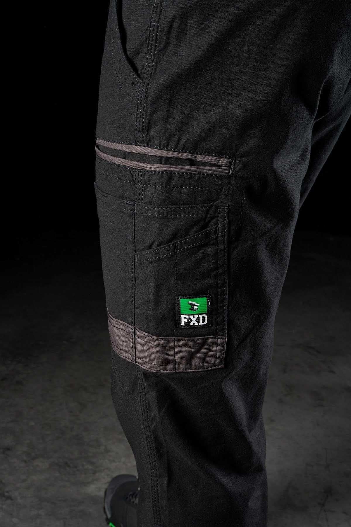 Close-up of a person's leg wearing black FXD WP-4 Stretch Cuffed Work Pants with green and grey details. Made from stretch cotton canvas, these pants feature multiple pockets, including one with a prominently displayed FXD logo. The image focuses on the side of the leg, highlighting the durable fabric and stitching.