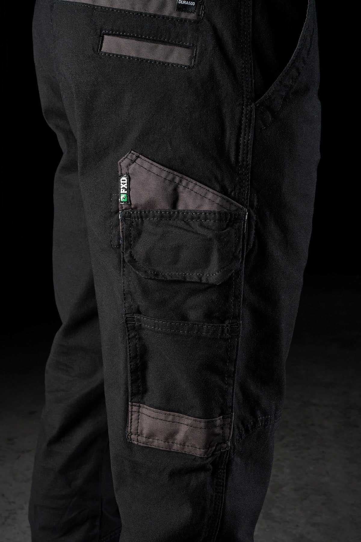 Close-up of a person wearing FXD WP-4 Stretch Cuffed Work Pants made from stretch cotton canvas with multiple pockets. The pants feature a combination of black fabric and dark grayish-brown DURA500 Advanced Nylon reinforcements on the pockets and knees. One pocket has a small label with green, white, and black colors. The background is dark.