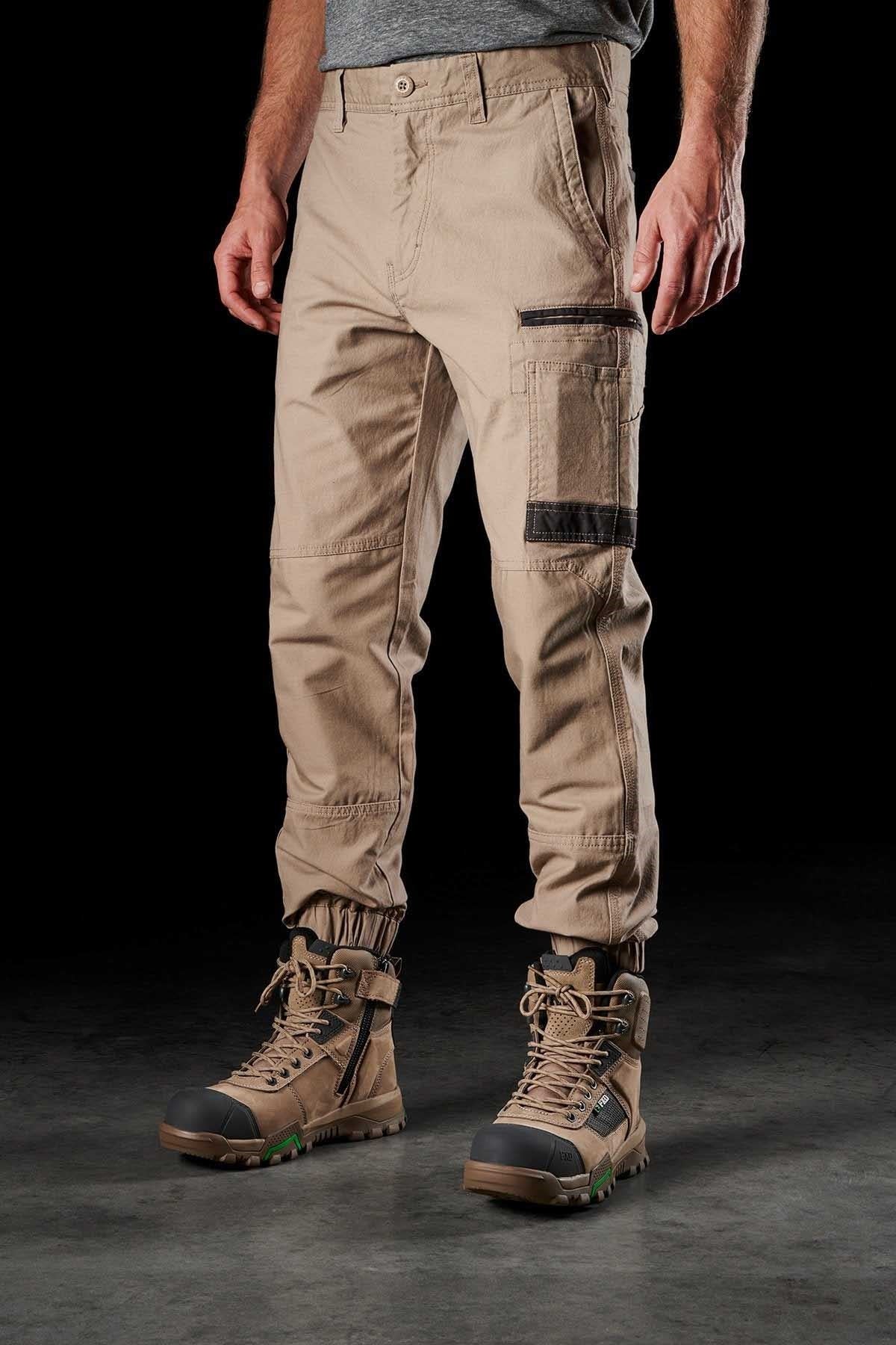 A man wearing tan FXD WP-4 Stretch Cuffed Work Pants, expertly crafted from durable stretch cotton canvas, and paired with sturdy brown lace-up work boots stands on a dark surface against a black background. While his upper torso isn't visible, the pants exude durability, making them perfect for manual labor.