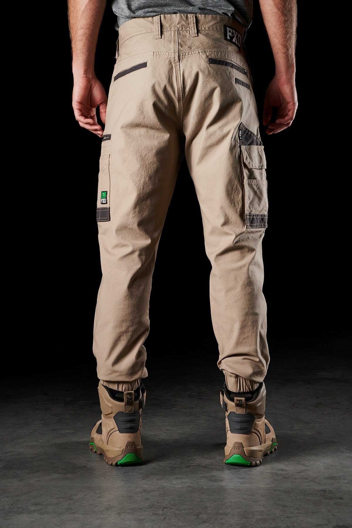 A person is standing with their back to the camera, wearing FXD WP-4 Stretch Cuffed Work Pants in beige and tan work boots with green soles. The pants, made from stretch cotton canvas, feature multiple pockets and loops against a dark background.