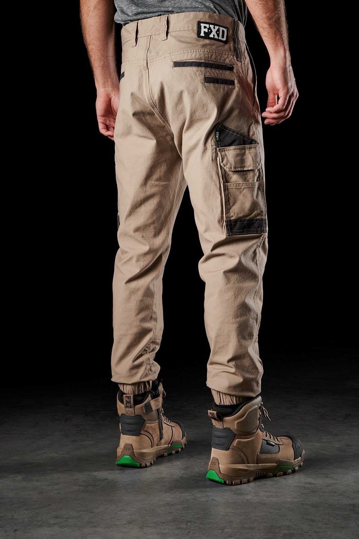 A person stands facing away, wearing FXD WP-4 Stretch Cuffed Work Pants in light brown stretch cotton canvas with multiple pockets and black accents. The pants have an "FXD" logo on the back waistband. The person is also wearing rugged tan and black work boots with green soles. The background is dark.
