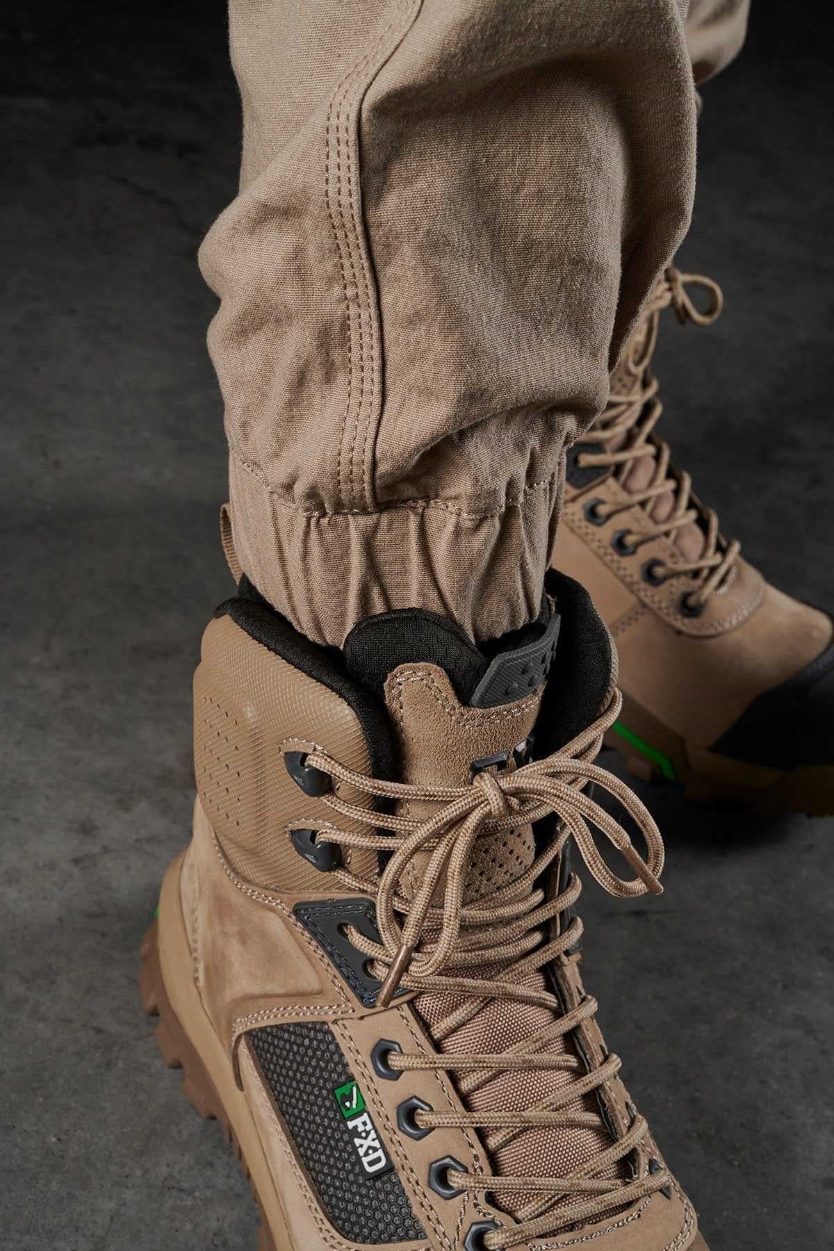 Close-up of a person wearing tan-colored, high-top hiking boots with laces tied up and FXD WP-4 Stretch Cuffed Work Pants made from DURA500™ Advanced Nylon And Polyurethane Canvas. The background is a dark, textured surface.