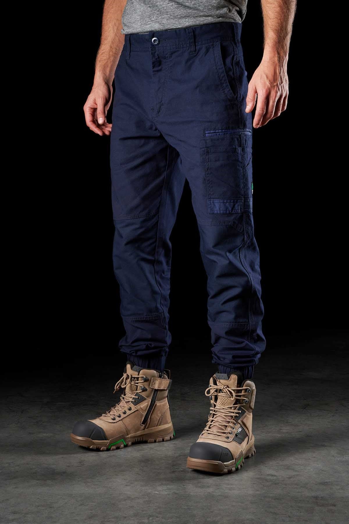 Man in a gray t-shirt showcases FXD WP-4 Stretch Cuffed Work Pants made from stretch cotton canvas, featuring multiple pockets and drawstring hems, paired with tan work boots. The outfit, enhanced with DURA500™ reinforcement, is displayed against a plain black background.