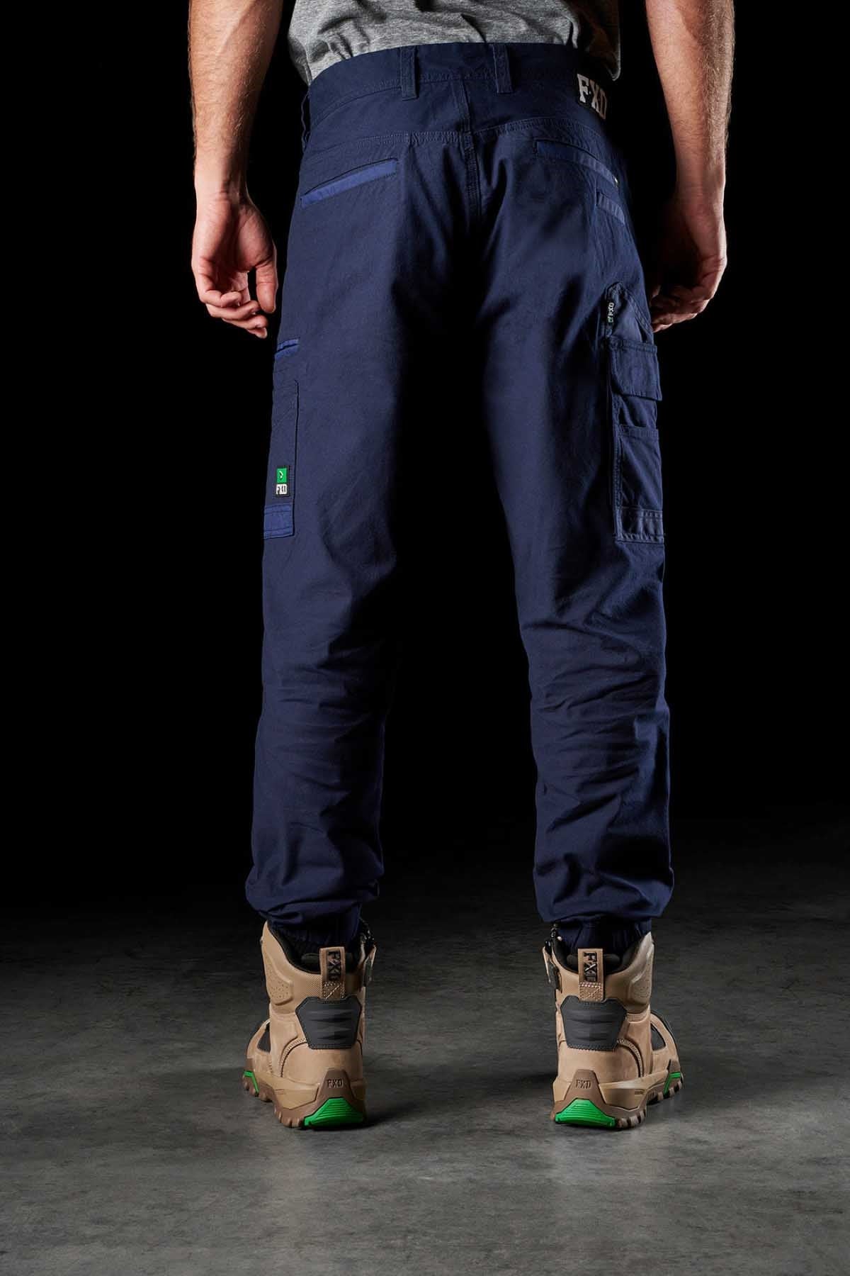 A person wearing dark blue FXD WP-4 Stretch Cuffed Work Pants and tan work boots stands with their back to the camera against a black background. The stretch cotton canvas pants, reinforced with DURA500™, have multiple pockets, including a large one on the right thigh, and the boots feature green accents on the soles.